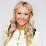 Smiling woman with long, wavy blonde hair, wearing a white blouse, poses in front of a plain light background.