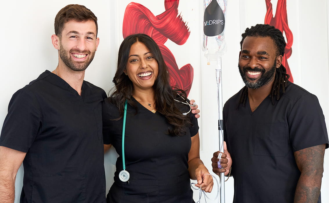 Three healthcare professionals in black scrubs smile together in a brightly lit room, with medical equipment and abstract red art on the wall behind them. One holds a stethoscope, another holds an IV drip.
