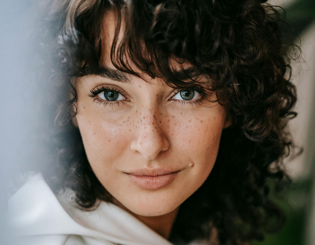 A person with curly dark hair, green eyes, freckles, and light skin looks directly at the camera, wearing a white top.